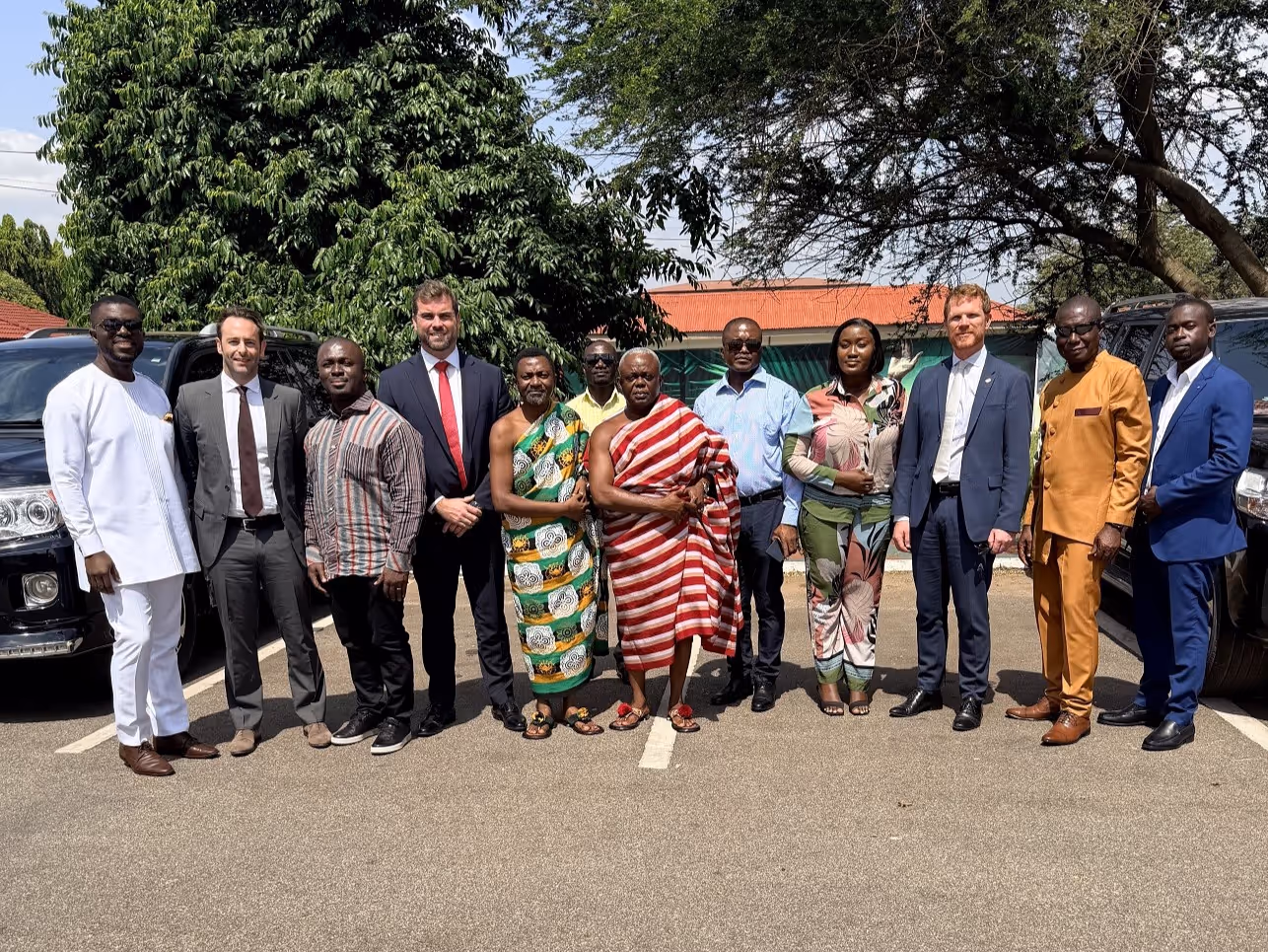 Thirteen people standing outdoors on a parking lot, dressed in a mix of traditional African attire and business suits, with trees and vehicles in the background.